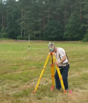 Zdjecie geodety dokonującego pomiaru z wykorzystaniem instrumentów geodezyjnych na otwartym terenie pod lasem.